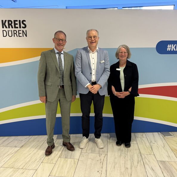 Das Bild zeigt zwei Männer und eine Frau vor einer Stellwand des Kreises Düren